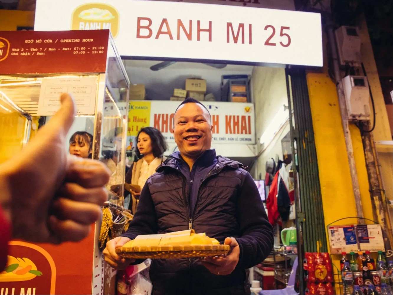 Person holding food outside a shop with a 'Banh Mi 25' sign; a thumbs-up gesture in the foreground.