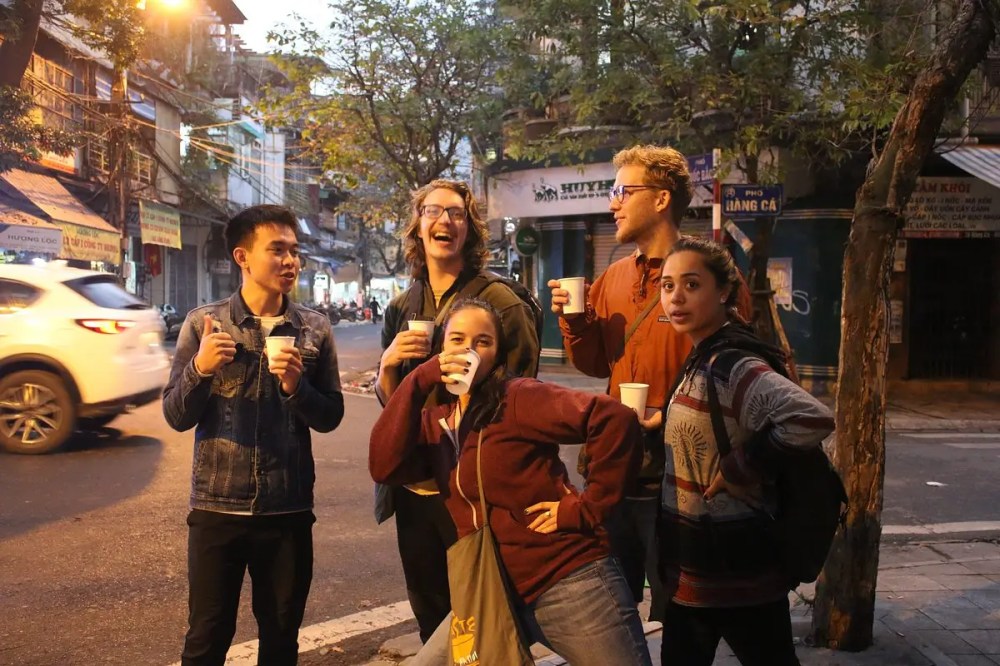 Five people standing on a street, holding cups, with a car passing by.